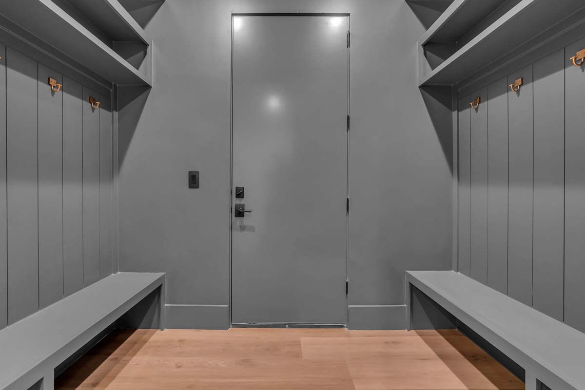 A gray locker room with wooden benches along the walls. The room features coat hooks and a closed gray door at the center, with wooden floors contrasting the overall gray interior.