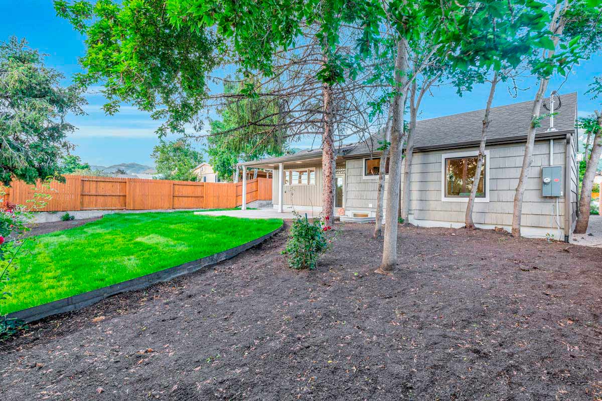 A backyard with a well-maintained lawn bordered by a black edge, several large trees, a wooden fence, and a gray house on the right side. The sky is clear and blue.