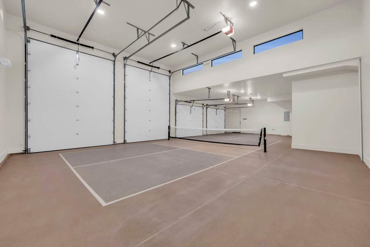 A spacious indoor room with light brown flooring and white walls, featuring a small tennis court setup with a net. The ceiling has bright lights, and there are several large, closed garage doors with windows above them.