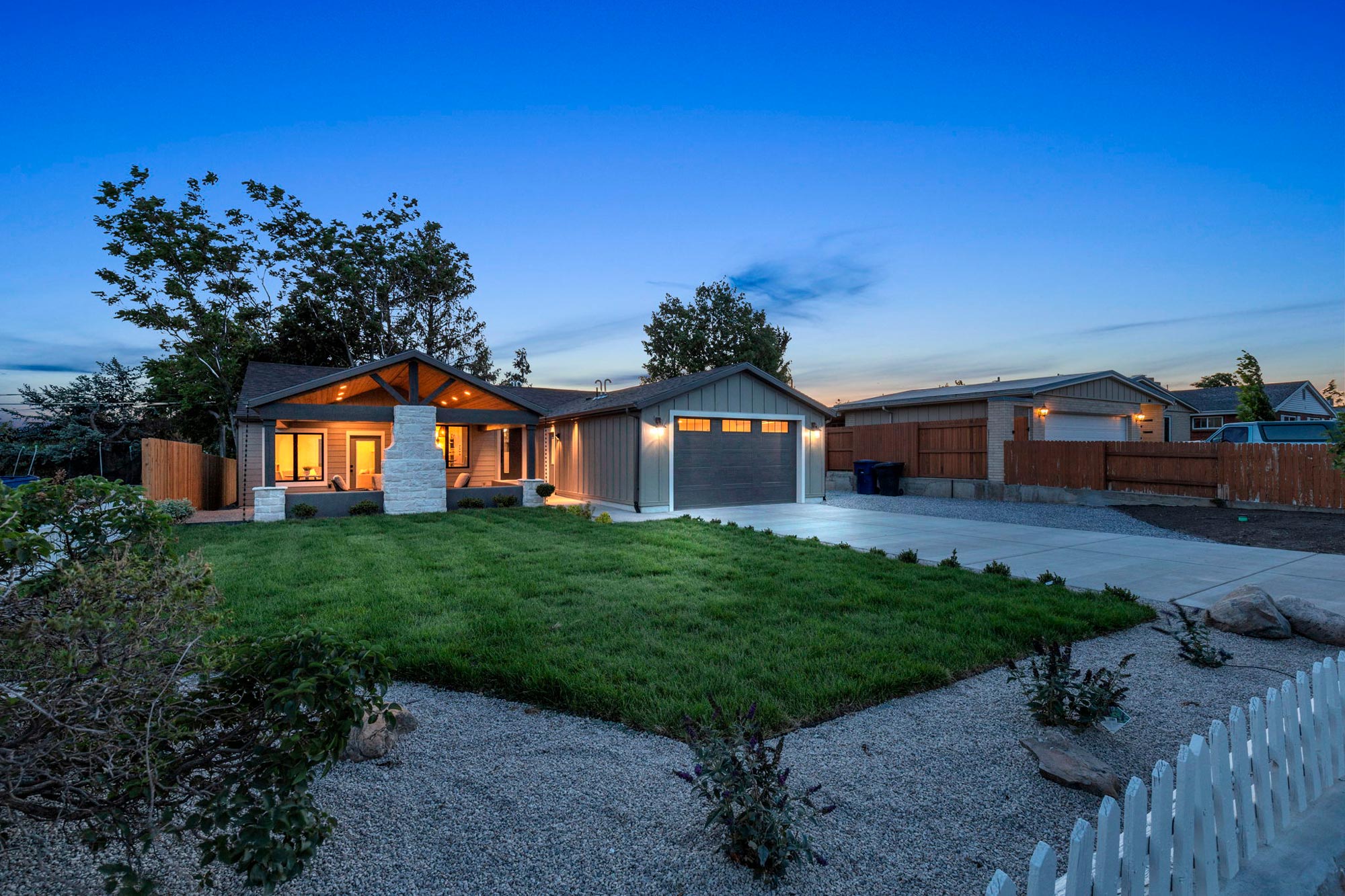 A single-story house at dusk with a large front lawn and a driveway leading to a two-car garage. The home features a welcoming porch with warm lights. A white picket fence and gravel path border the grass, with trees in the background under a clear sky.