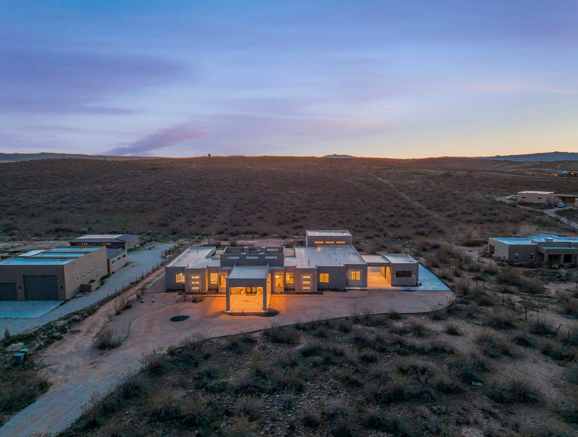 Modern house in a desert landscape at dusk, with warm lights glowing from the windows. The sky is a mix of purples and oranges as the sun sets, creating a tranquil atmosphere. A winding path leads to the house surrounded by sparse vegetation.