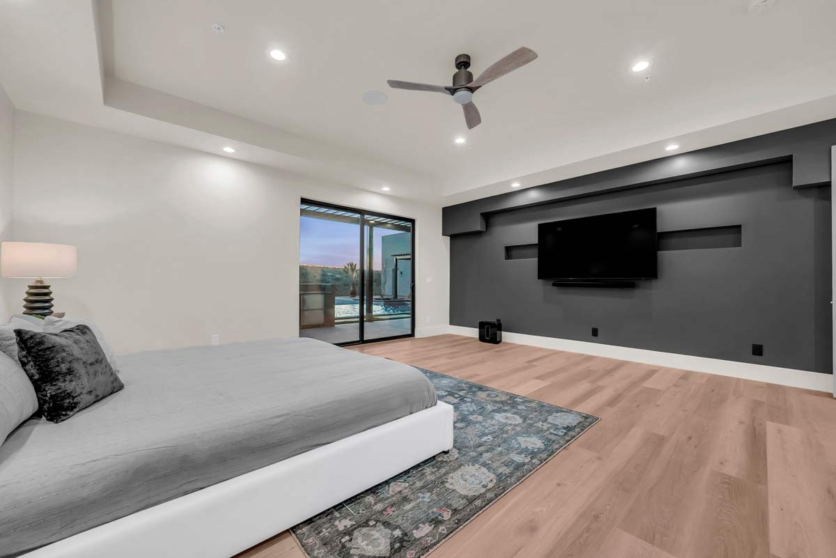 Modern bedroom with a large bed and gray accent wall featuring a mounted TV. A ceiling fan hangs above, and glass doors lead to a patio with a pool view. Light wood flooring and a patterned rug complete the contemporary design.