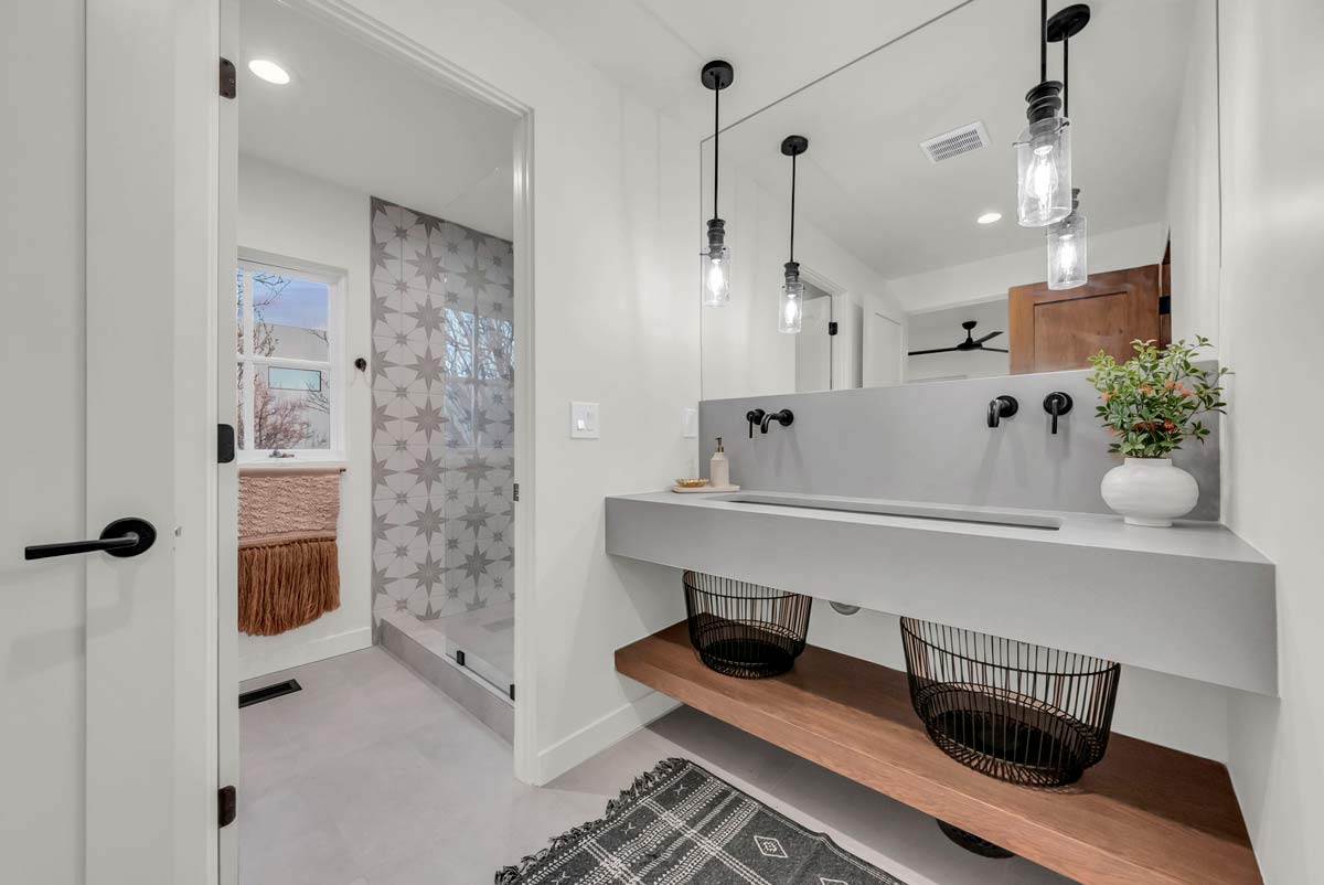 Modern bathroom with a minimalist design features a dual-sink vanity with hanging black wire baskets underneath, industrial-style pendant lights, a walk-in shower with patterned tiles, and a decorative plant on the counter.