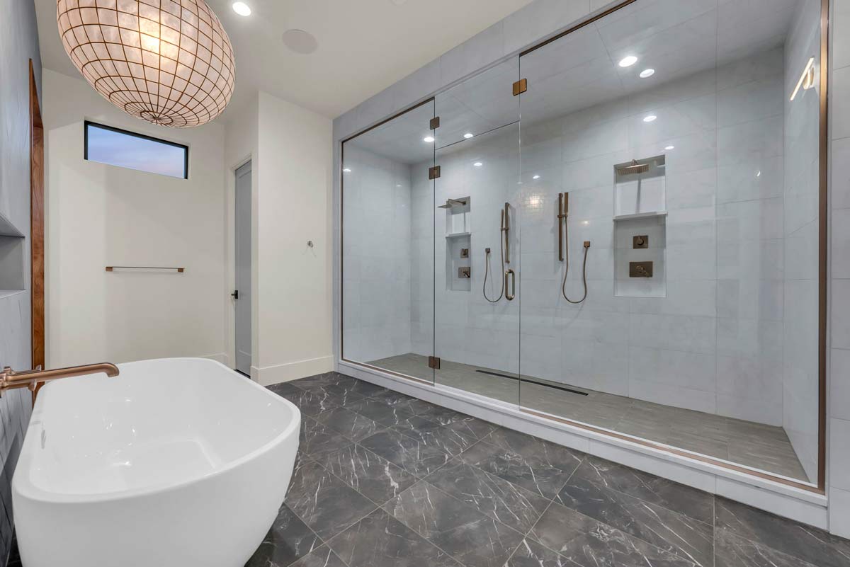 Modern bathroom with gray marble floor, a white freestanding bathtub, and a large glass-enclosed shower area featuring dual showerheads and niches. A round pendant light hangs above, and a small window allows natural light in.