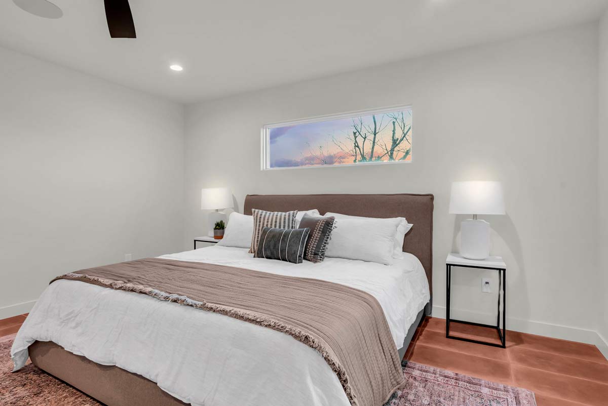 A modern bedroom features a large bed with white and beige bedding. Two bedside tables with lamps and a potted plant complement the space. A narrow horizontal window above the bed reveals a sunset sky with bare tree branches.