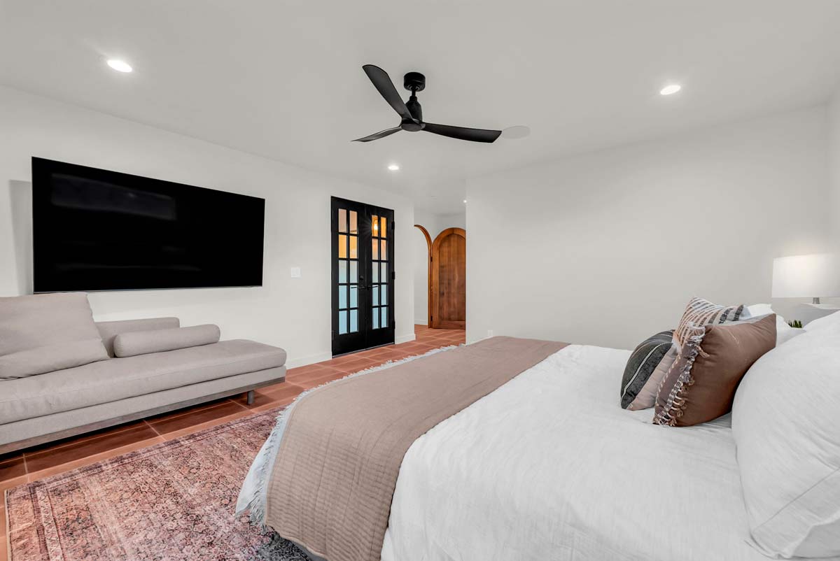 Minimalist bedroom with a large bed featuring white and beige bedding. A modern ceiling fan, flat-screen TV, and a beige sofa are visible. Theres a patterned rug on a wooden floor and black double doors leading to another area.