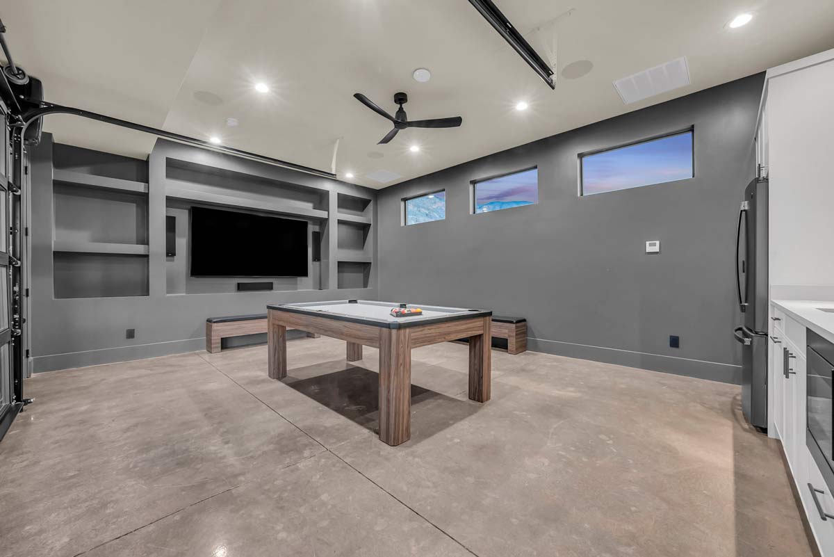 Modern game room with a sleek pool table centered on a concrete floor. The walls are dark gray with built-in shelves and a large flat-screen TV. Three rectangular windows line the upper wall, beneath a ceiling fan with recessed lighting.