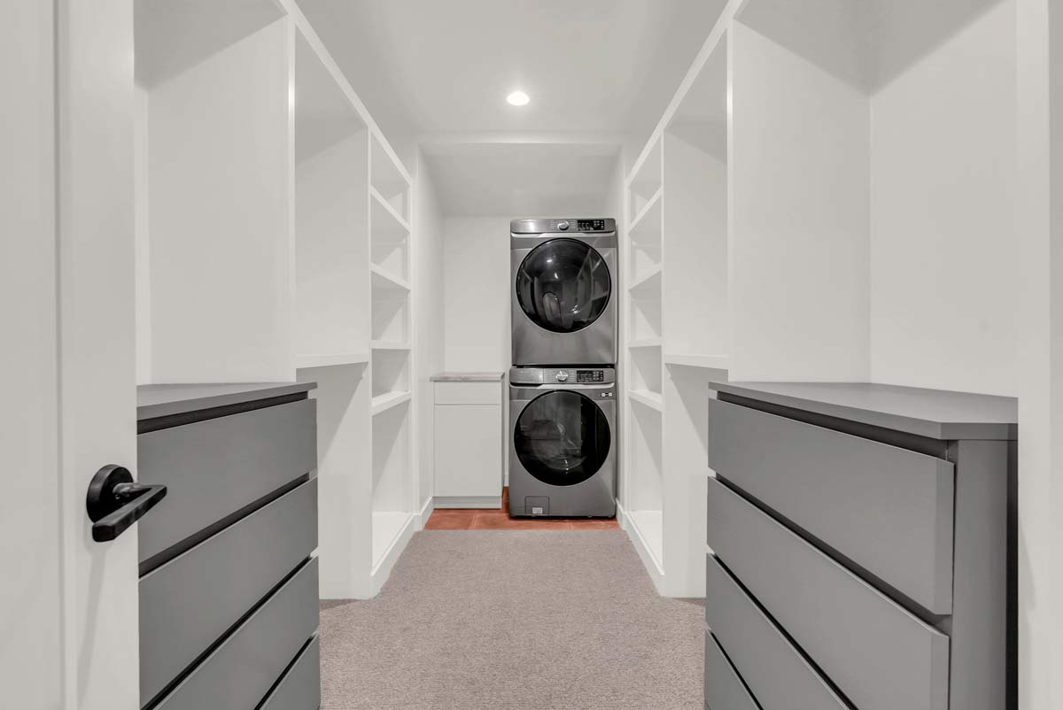 Modern laundry room with stacked washer and dryer at the far end. Open shelving lines both sides, and two gray dressers are positioned along the walls. Carpeted floor and white walls create a bright, clean look.