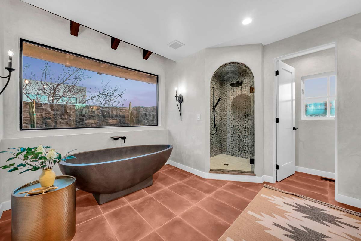 Modern bathroom featuring a freestanding dark bathtub, arched stone-tiled shower, large window with outdoor view, terracotta floor tiles, and a geometric-patterned rug. A small table with a plant sits next to the tub.
