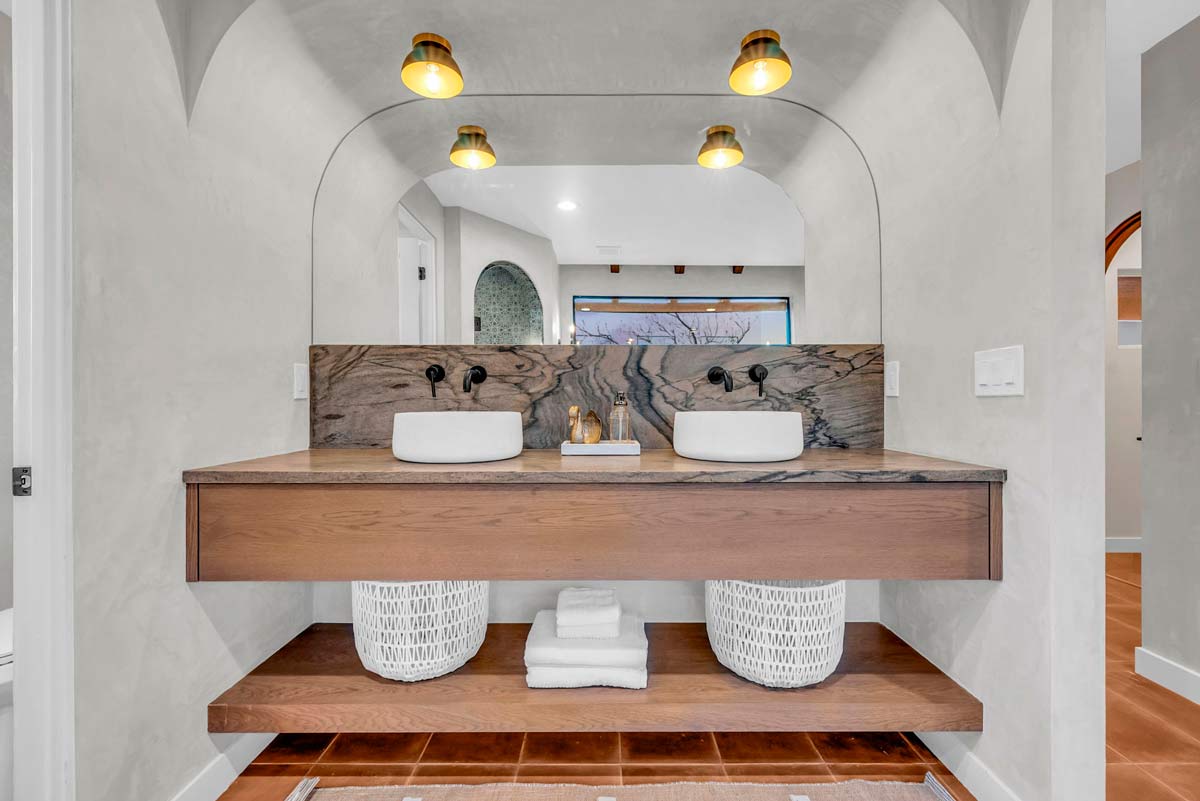 Modern bathroom with two vessel sinks on a wood vanity, paired with rounded mirrors and stylish black fixtures. Underneath, white baskets and neatly folded towels add a touch of elegance. Warm lighting and an archway enhance the sophisticated look.