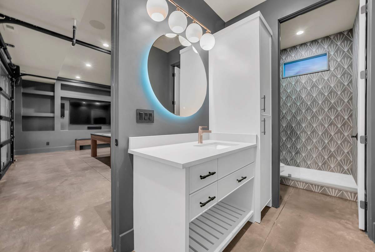 Modern bathroom with a white vanity, round mirror, and three globe lights. Geometric patterned wallpaper adorns the shower area with a small window. To the left, a corridor leads to a room with a pool table and large TV.