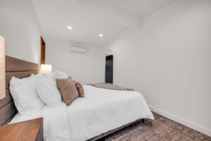 Cozy bedroom with a large bed covered in white linens and several pillows. The room features a wooden headboard, bedside table with a lamp, and a patterned rug. Walls are plain with a visible door and an air conditioning unit above.