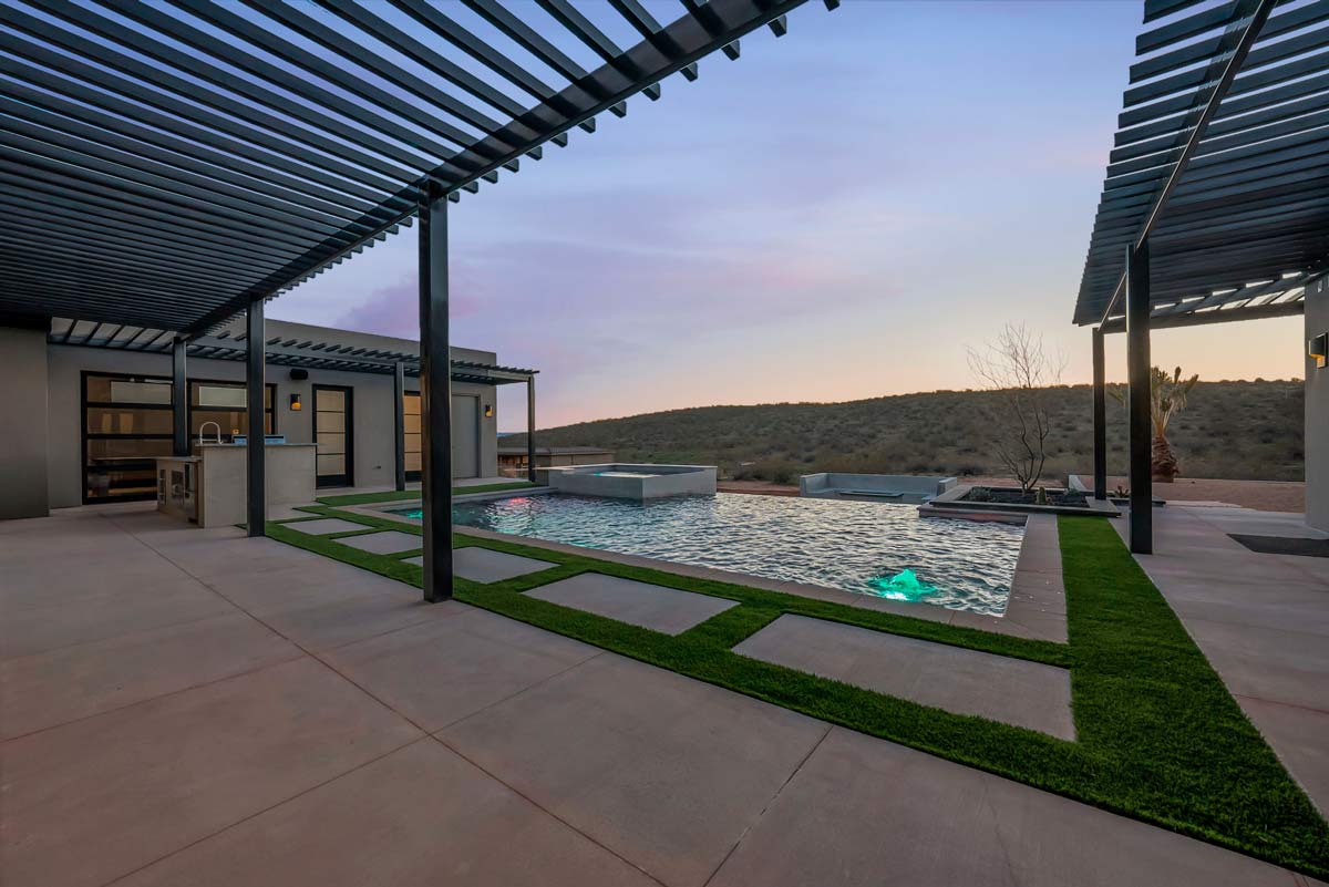Modern outdoor space featuring a sleek swimming pool with embedded pool lights. The area is surrounded by smooth concrete and strips of grass, with pergolas providing shade. The backdrop reveals a desert landscape under a twilight sky.