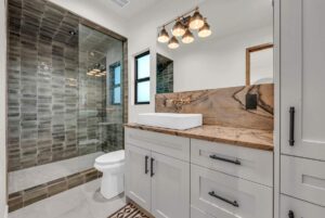 Modern bathroom with a white vanity, marble countertop, and square sink. A large mirror is above, with stylish light fixtures. The shower features dark green tiles and a glass door. A toilet and window complete the space.