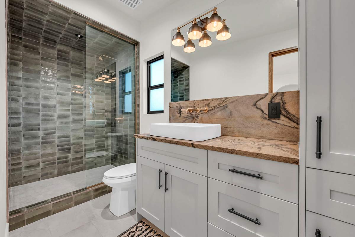 Modern bathroom with a white vanity, marble countertop, and square sink. A large mirror is above, with stylish light fixtures. The shower features dark green tiles and a glass door. A toilet and window complete the space.
