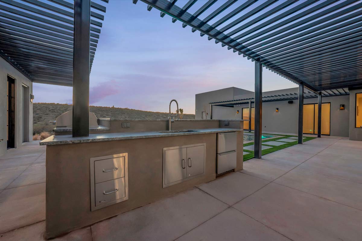 Modern outdoor patio with a built-in kitchen featuring a sink, grill, and storage. The area is covered by a sleek, slatted pergola. There is a small grassy patch and a pool in the background, surrounded by concrete flooring.