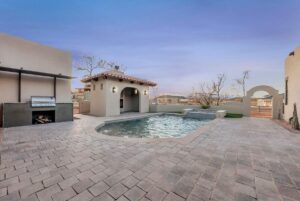 Outdoor patio with a small swimming pool surrounded by stone tiles. Theres a barbecue grill on the left and a small structure with a tile roof. Bushes and desert landscape visible in the background under a clear sky.