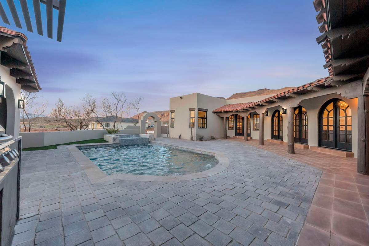 A spacious courtyard with a small pool, surrounded by a rustic-style building with arched windows and doors. The sky is clear, showcasing a sunset. Hills are visible in the background, and the paving is neatly arranged around the pool area.