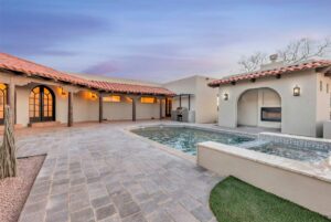 A spacious courtyard features a rectangular pool surrounded by a tiled patio. The area is enclosed by a house with a terracotta roof and arched windows. Warm lights illuminate the space, and a serene evening sky is in the background.
