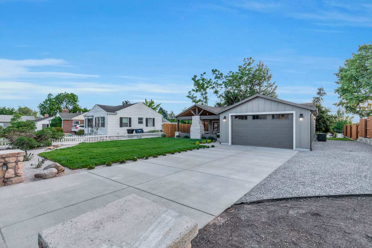 Suburban neighborhood with a modern gray house featuring a large garage, adjacent to a white house with a picket fence. Well-maintained lawns and a paved driveway are visible under a clear blue sky.