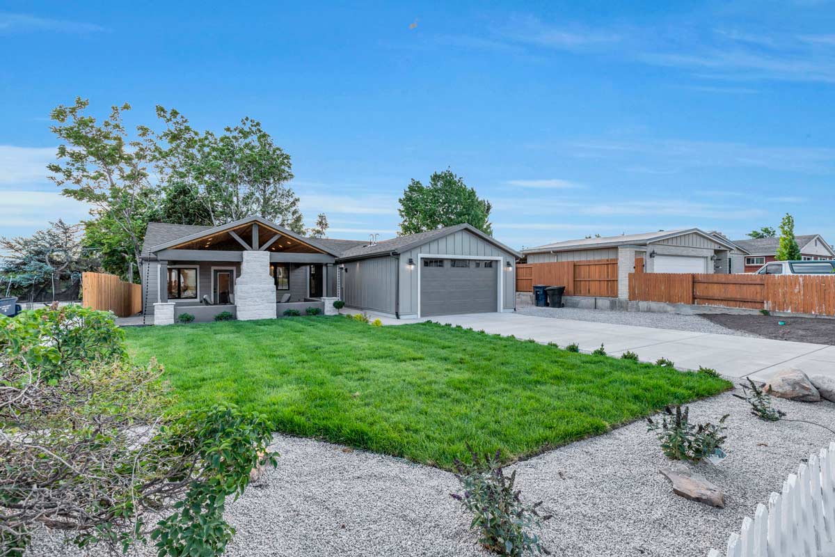 A modern suburban house with a clean, angular design sits behind a lush green lawn. The structure features a gray exterior with a prominent covered porch. The driveway and manicured landscape are visible under a clear blue sky.