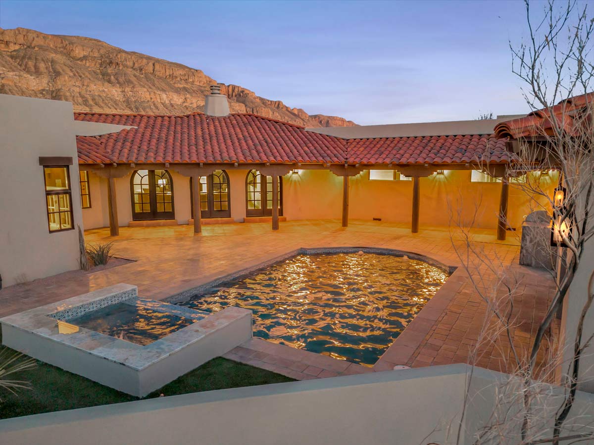 A courtyard with a pool and jacuzzi surrounded by a Spanish-style house with red tile roof. Large windows line the house. Mountains are visible in the background during sunset, casting a warm glow over the scene.