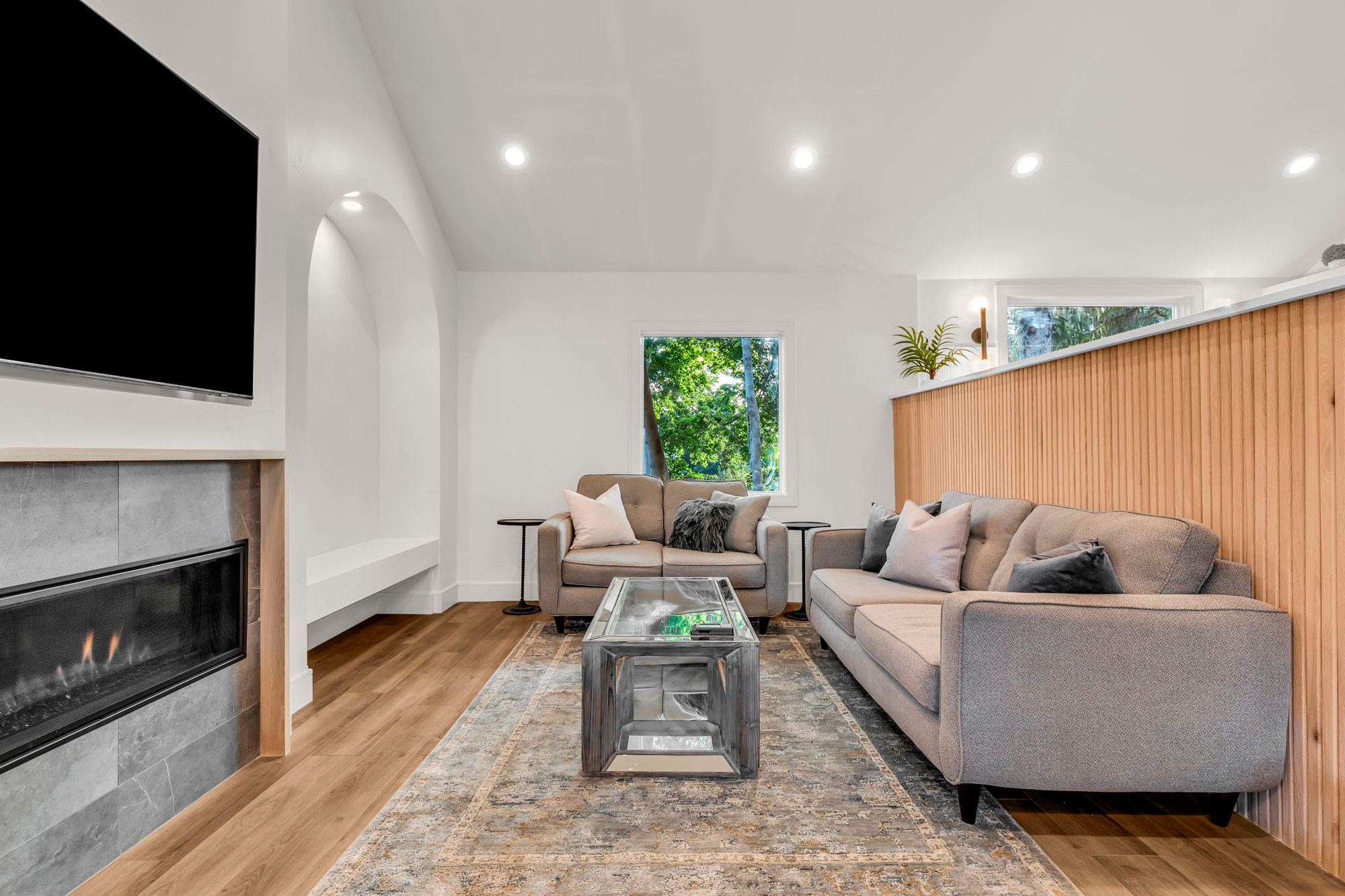 A modern living room with a gray sofa, armchairs, and a glass coffee table on a patterned rug. A flat-screen TV is mounted above a sleek fireplace. A large window lets in natural light, highlighting the wood paneling and minimalist decor.