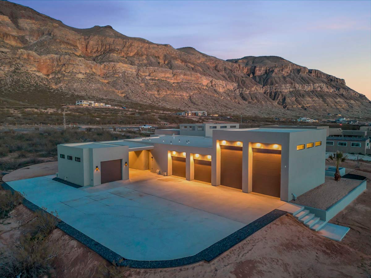Modern house with a flat roof and large garage doors, situated in a desert landscape at twilight. The building features minimalist design with large windows, surrounded by rocky hills in the background and a paved driveway in the foreground.
