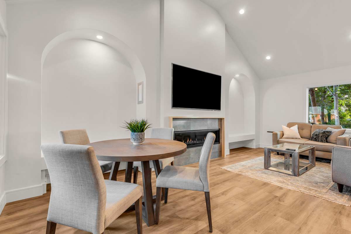 A modern living area with a round wooden dining table and four upholstered chairs. Theres a wall-mounted TV above a fireplace, a cozy seating area with a sofa and coffee table, and large windows overlooking greenery. Wood flooring throughout.