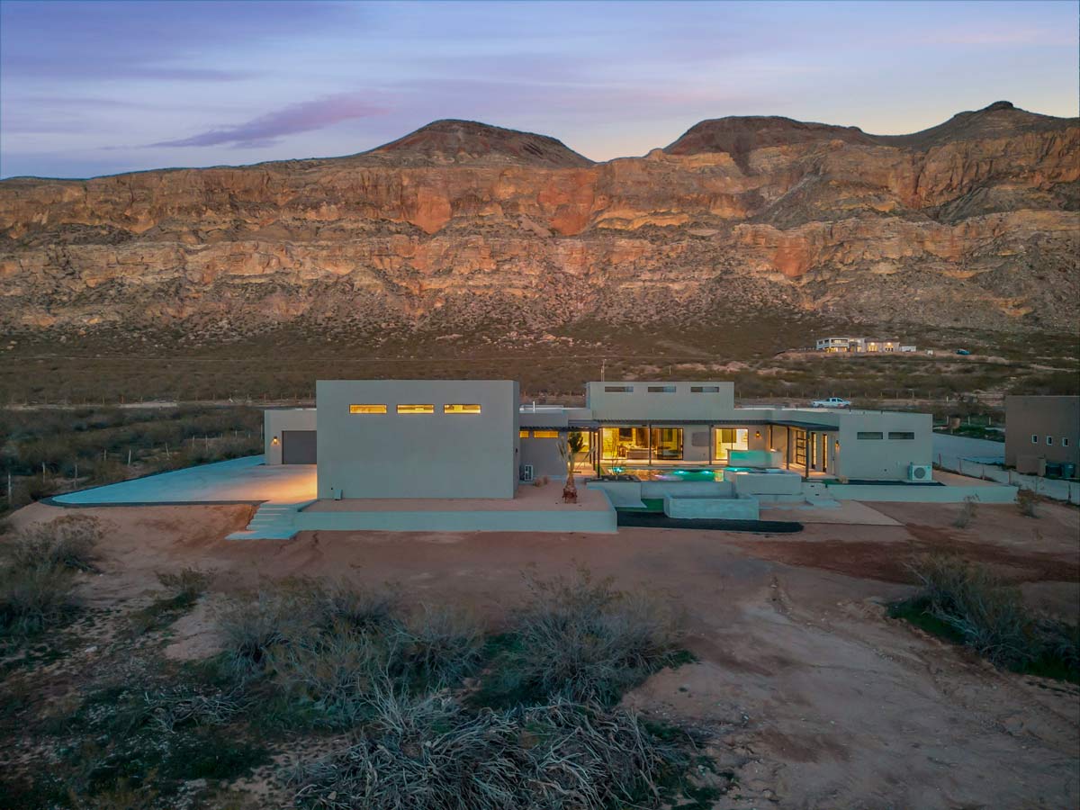 A modern, low-rise house with a minimalist design is nestled in a desert landscape. The home features large windows and a swimming pool, set against striking, rocky hills under a dusky sky. Sparse vegetation surrounds the area.