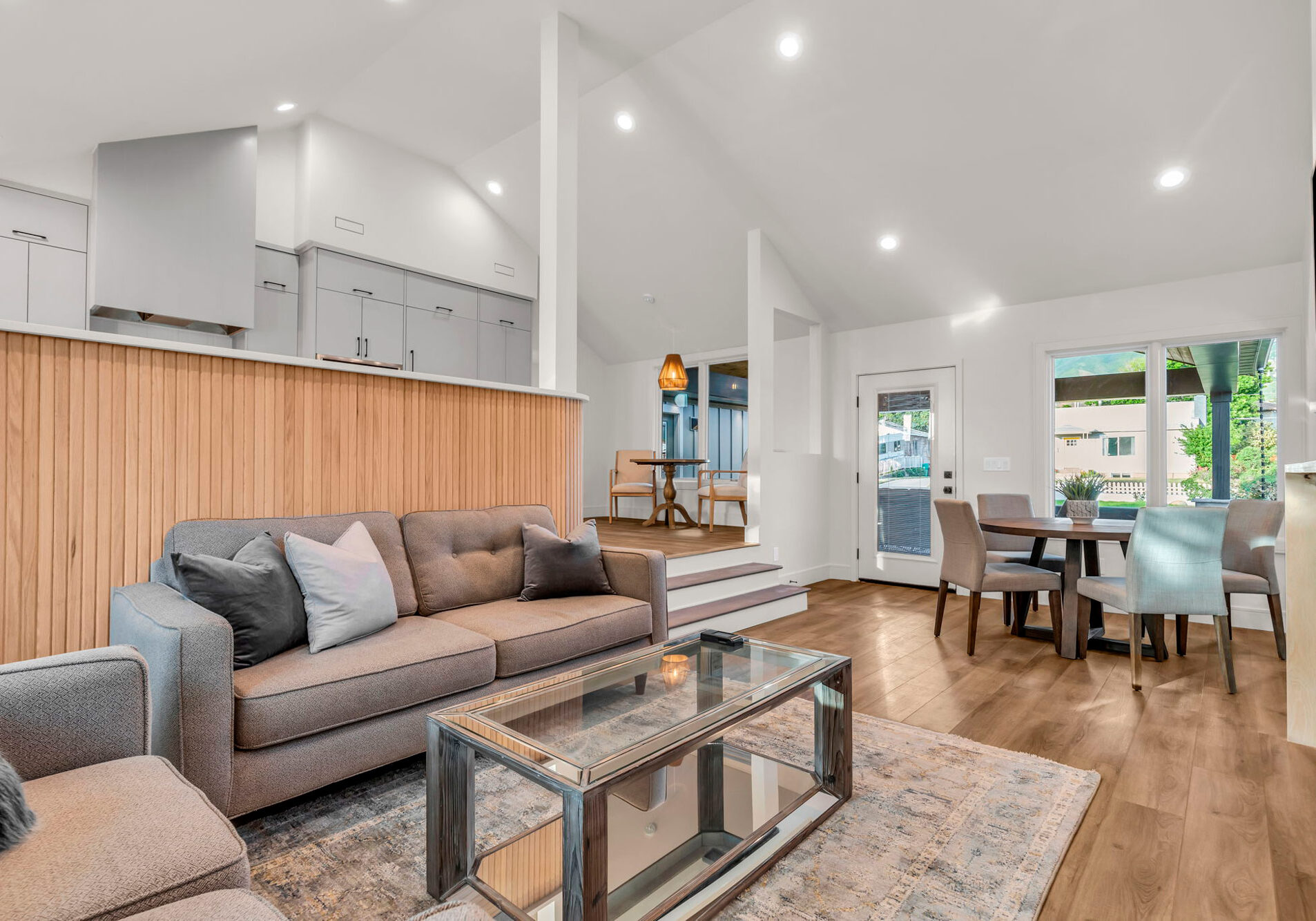 Modern open-concept living room and kitchen with hardwood floors. The space features a gray sofa, glass coffee table, wooden accent wall, and dining table by a large window. Bright lighting and a blend of contemporary decor create a welcoming atmosphere.